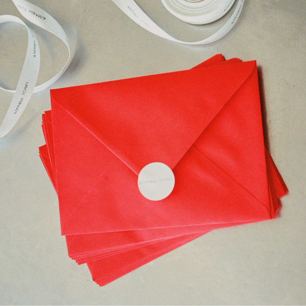 A stack of bright red envelopes with white circular seals, arranged on a light beige surface with a white ribbon partially visible in the background.